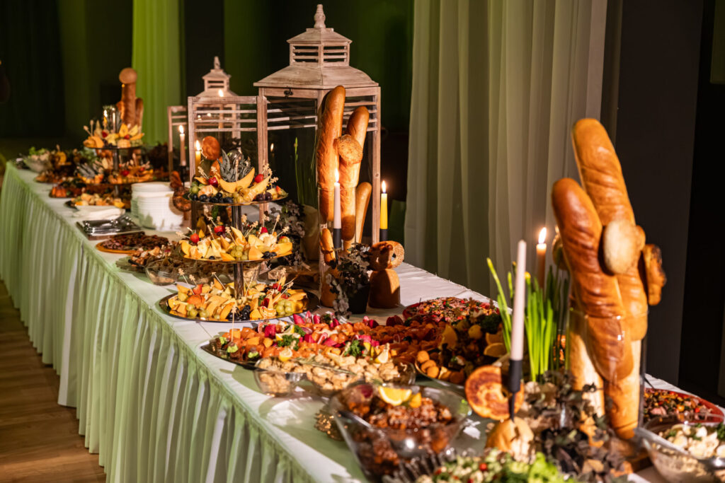 A grand buffet table adorned with bread centerpieces, candles, and wooden lanterns, filled with colorful fruit, hors d'oeuvres, and gourmet dishes under elegant lighting
