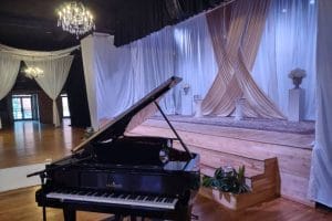A grand piano is positioned in front of a stage with draped white and beige curtains, and vases of flowers on pedestals. Two chandeliers are visible, and wooden flooring covers the venue—creating exactly what guests want from a wedding reception.