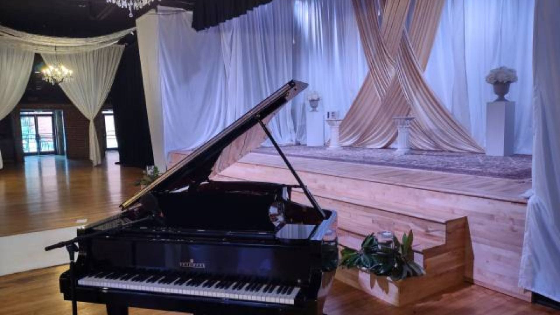 A grand piano is placed near a stage adorned with white and gold drapes and floral arrangements in an elegantly decorated event hall, offering exactly what guests want from a wedding reception.