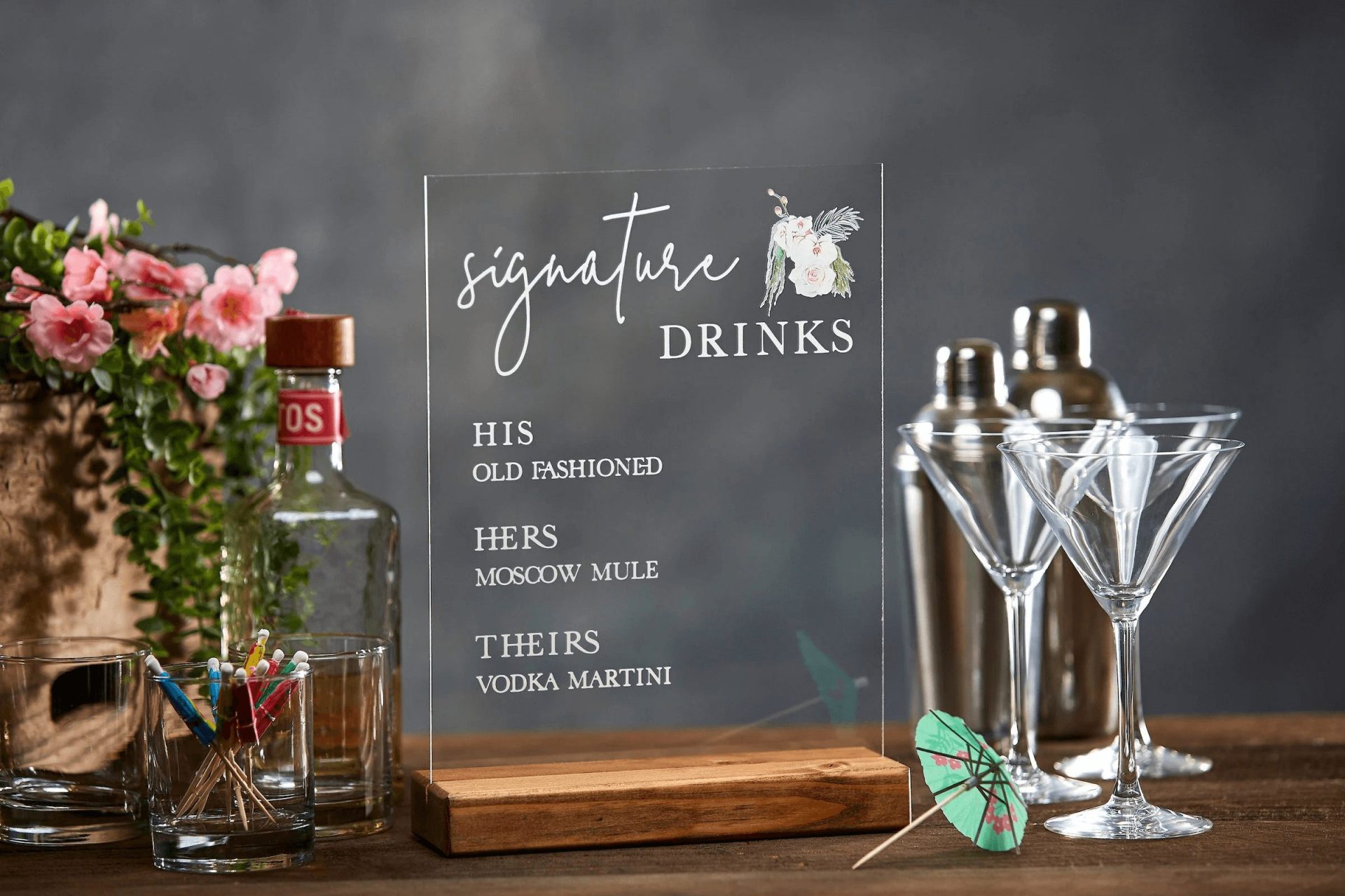 A sign titled "Signature Drinks" lists "His: Old Fashioned," "Hers: Moscow Mule," and "Theirs: Vodka Martini." It stands beside bottles, cocktail shakers, and martini glasses on a table.